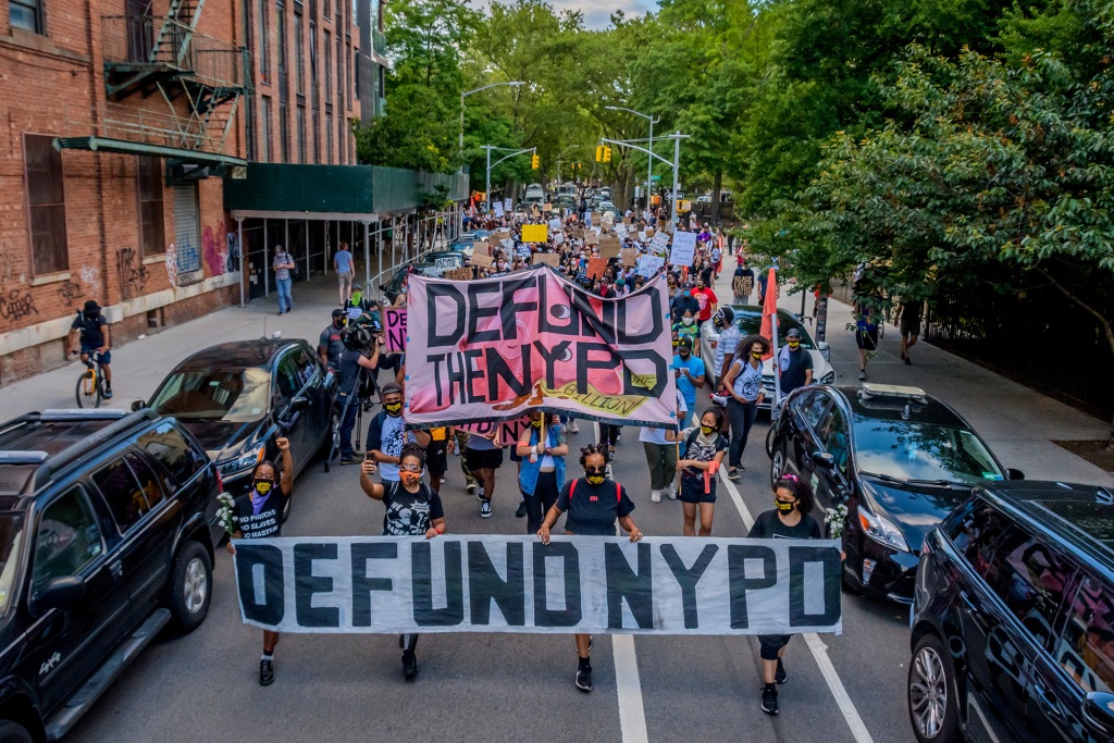 A large crowd of people stretches down a city roadway holdings signs and banners that read "Defund the NYPD." 
