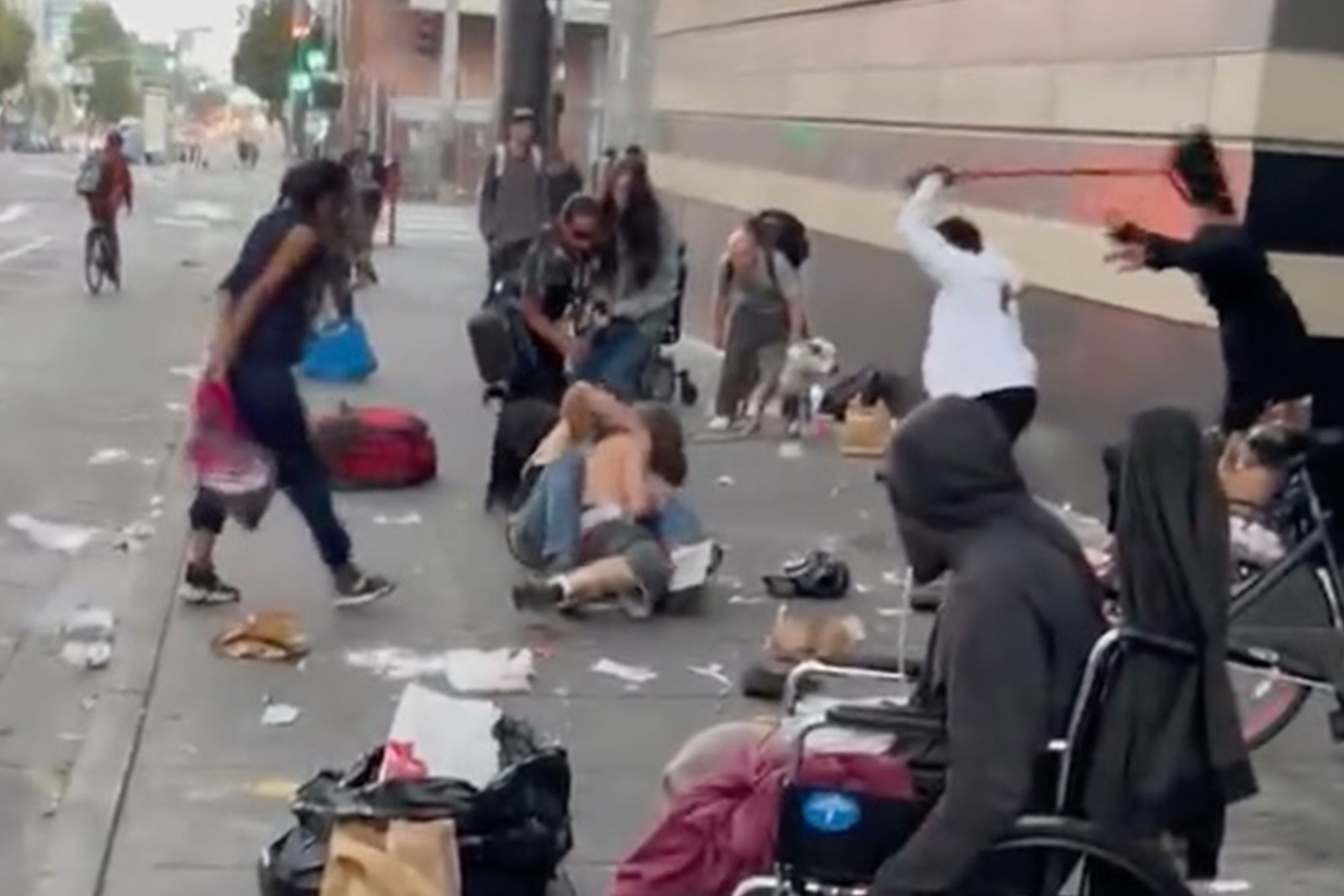 Two homeless men wrestle on the ground, while a third whacks one of them with a broom.